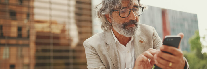 Mature businessman with beard in eyeglasses wearing gray jacket is using cell phone, Panorama....