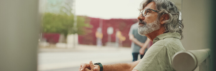 Thoughtful, middle-aged man with gray hair and beard, wearing casual clothes, sits at public...