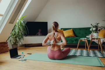 Poster Back view of woman is sitting in a lotus position on a yoga mat in a living room © Daria