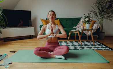 A woman is leisurely sitting crosslegged on a yoga mat in a living room