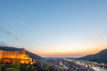 View over an old town with a castle or palace rune in the evening at sunset. This place is located in a river valley of the Neckar, surrounded by hills. Heidelberg, Baden-Württemberg, Germany - obrazy, fototapety, plakaty
