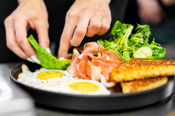 A chef garnishes a gourmet breakfast plate with fresh green salad, showcasing vibrant colors and textures of eggs, meat, and vegetables