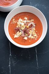 Spanish salmorejo served with prosciutto and grated egg in a white bowl, vertical shot on a black marble background, elevated view