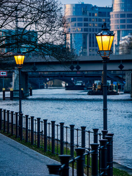 Portrait of Bellevue Ufer waterfront offers a view of Spree river, adorned with glowing street lamps, leading to sunlit office buildings in Berlin Moabit, framed by bridges including Moabit bridge.