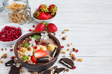 Chocolate smoothie bowl topped with oat granola, chocolate cookie with cream, banana, strawberries, pomegranate and spring flowers on white wooden table. Healthy vegan protein food for breakfast