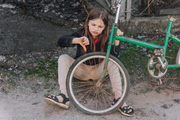 A small beautiful sad teenage girl, a dissatisfied upset child, showing a dislike, sits near an old bicycle with a broken, punctured wheel tire outdoors. Photography, portrait.