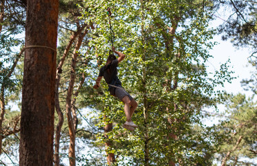 A man in a black T-shirt and shorts is engaged in a tightrope walk in the forest
