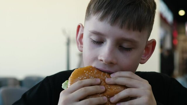 A 9-year-old Caucasian boy eats a delicious burger while in a fast food cafe. a child has lunch with a big burger in the school cafeteria. fast food and teenager