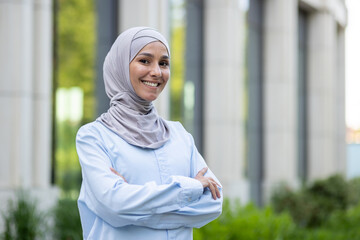 A professional Muslim woman in a hijab stands confidently outdoors with her arms crossed, exuding...