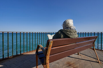 Mujer senior lee tranquila y calmada frente a la playa en el mar mediterráneo. - obrazy, fototapety, plakaty
