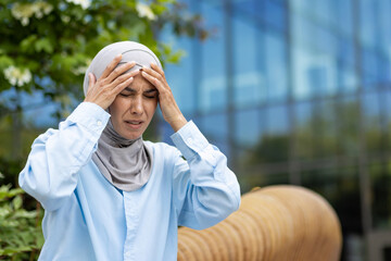 A professional woman in a hijab looks stressed and holds her head, indicating a headache or...