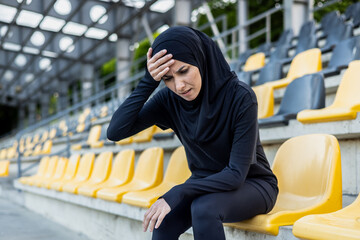A tired female athlete in hijab sitting on stadium seats, showing signs of fatigue and exhaustion...