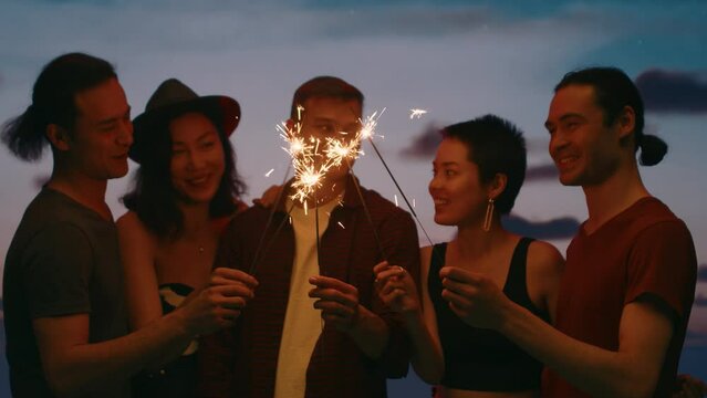 Young friends with sparklers talking and smiling close up standing in a circle at an outdoor party with a spectacular dusk sky in a backdrop. Concept of carefree summer leisure