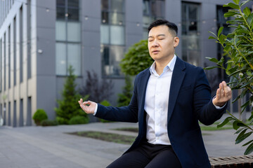 Serious Asian male businessman sitting on bench near working office in lotus position and...