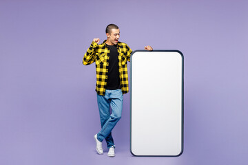 Full body young happy middle eastern man wear yellow shirt casual clothes big huge blank screen mobile cell phone smartphone with workspace area do winner gesture isolated on plain purple background.