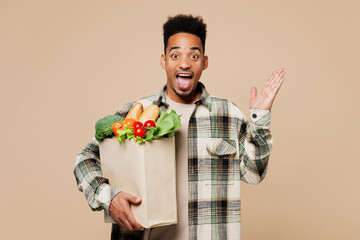 Young shocked surprised man wear grey shirt hold paper bag for takeaway mock up with food products spread hand isolated on plain pastel light beige background Delivery service from shop or restaurant