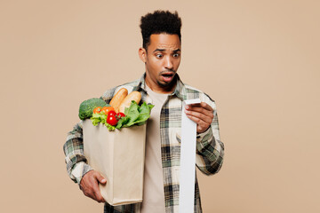 Young sad mad man in grey shirt hold paper bag for takeaway mock up with food products read cash bill receipt isolated on plain pastel light beige background Delivery service from shop or restaurant