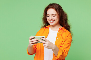 Young happy gambling ginger woman wear orange shirt white t-shirt casual clothes use play racing app on mobile cell phone hold gadget smartphone for pc video games isolated on plain green background.