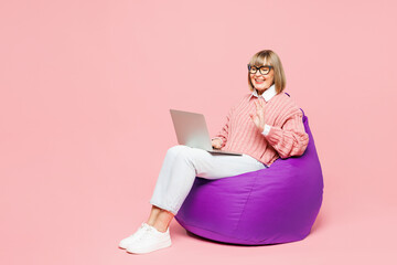 Full body elderly IT woman 50s years old wear sweater shirt casual clothes glasses sit in bag chair hold use work on laptop pc computer waving hand isolated on plain pink background Lifestyle concept - obrazy, fototapety, plakaty