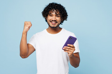 Young happy Indian man he wear white t-shirt casual clothes hold in hand use mobile cell phone do winner gesture isolated on plain pastel light blue cyan background studio portrait. Lifestyle concept.