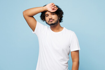 Young sad sick ill Indian man he wearing white t-shirt casual clothes put hand on forehead suffer from headache isolated on plain pastel light blue cyan background studio portrait. Lifestyle concept.