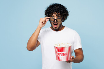 Young shocked amazed Indian man he wear white t-shirt casual clothes lower 3d glasses watch movie film hold bucket of popcorn in cinema isolated on plain pastel light blue background studio portrait.