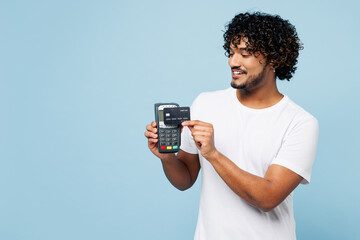 Young happy Indian man wears white t-shirt casual clothes hold wireless modern bank payment terminal to process acquire credit card isolated on plain pastel light blue background. Lifestyle concept.