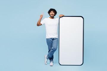 Full body young Indian man wear white t-shirt casual clothes big huge blank screen mobile cell phone smartphone with area point finger up isolated on plain pastel blue background. Lifestyle concept. - 778941281