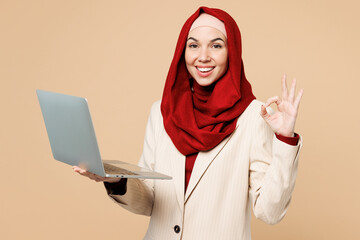 Young Arabian Asian Muslim IT woman wear red abaya hijab suit clothes hold use work on laptop pc computer show ok okay isolated on plain beige background. UAE middle eastern Islam religious concept.