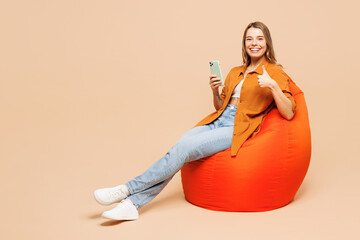 Full body young fun woman wear orange shirt casual clothes sit in bag chair hold in hand use mobile...