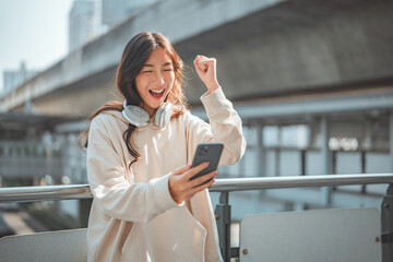 Excited asian woman wearing headphones reading on smartphone expressing happiness about great news, Female feeling like a winner while cheering for investment or banking app message in city