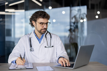 A professional male doctor in a lab coat using a laptop and writing notes on a clipboard in a...