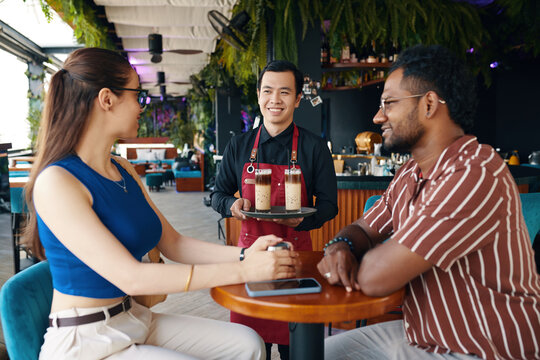 Smiling waiter bringing cold drinks to table of diverse couple