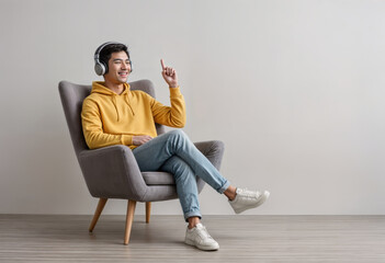 Joyful young Asian guy in wireless headphones sitting in armchair, having fun, listening to music, raising his arm, dancing to favorite song, enjoying cool soundtrack against white wall, empty space