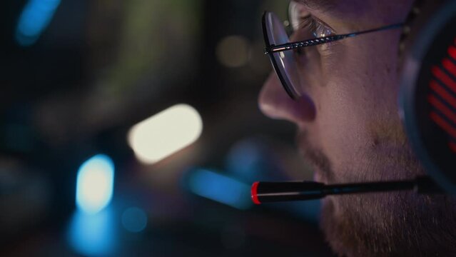 Gamer in glasses headset talks to team through mic close up. Adult bristly man plays video game at computer club. Esports tournament. Cyber sport championship. Modern gaming equipment earphones
