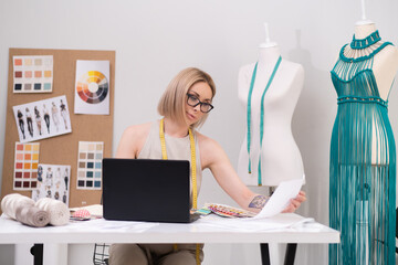 Blonde woman with glasses works on laptop at desk in tailoring workshop creative designer makes...