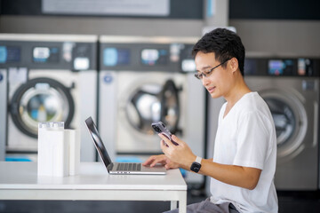 Young Asian man using laptop and smartphone doing his work while waiting for washing machine...