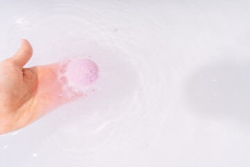 A woman's hand lowers a bath bomb into a bathtub of water.