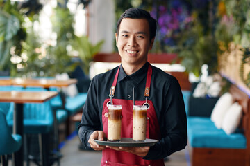 Portrait of smiling coffeeshop waiter holding tray with two glasses of iced coffee
