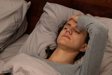 Healthcare concept of a tired, fatigued young woman lying in bed holding her head with her eyes closed; a sick female lying in bed with a hand on her forehead with closed eyes.