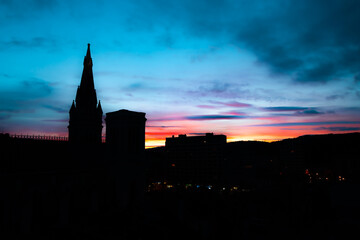 Buildings silhouetted against colourful, peaceful evening sky with blue, red, yellow sky and streetlights. Church steeple and tower against beautiful twilight sky. Sunset behind picturesque cathedral. - obrazy, fototapety, plakaty
