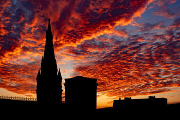 Obraz premium A dramatic sunset with fiery orange and yellow clouds with buildings silhouetted. Church steeple silhouette against a glorious, beautiful sunset with colourful clouds and blue and yellow sky..