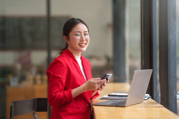 Asian businesswoman in a red suit is sitting at a table with a laptop and a cell phone