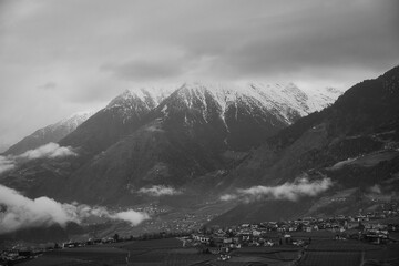 Schwarzweiss Aufnahme kleiner Orte in einem Tal mit massivem Bergpanorama mit Schnee auf den Gipfeln im Hintergrund - obrazy, fototapety, plakaty