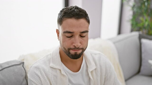 Upset young arab man frowning on home sofa, the nervous skeptic burdened with problems in his indoor lifestyle, grappily embracing a negative headspace.