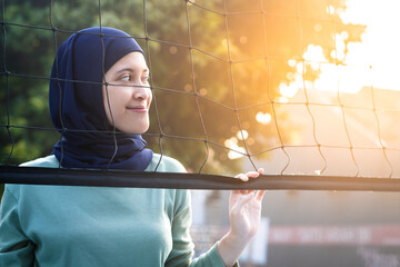 Muslim woman in hijab with brown eyes holding volleyball net in morning sunlight.