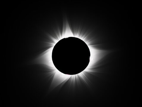 A black and white image of a solar eclipse, where the completely darkened Sun is surrounded by a bright corona, radiating into the dark space.