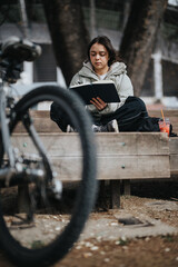 A serene scene captures a young woman deeply absorbed in reading a book, seated on a wooden bench beside her bicycle in a tranquil park setting.