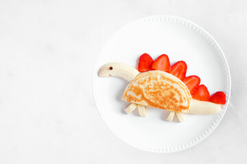 Fun child theme breakfast pancake in the shape of a dinosaur. Overhead view on a white marble background.