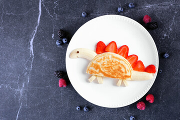 Fun child theme breakfast pancake in the shape of a dinosaur. Overhead view table scene on a dark stone background. - 779043426